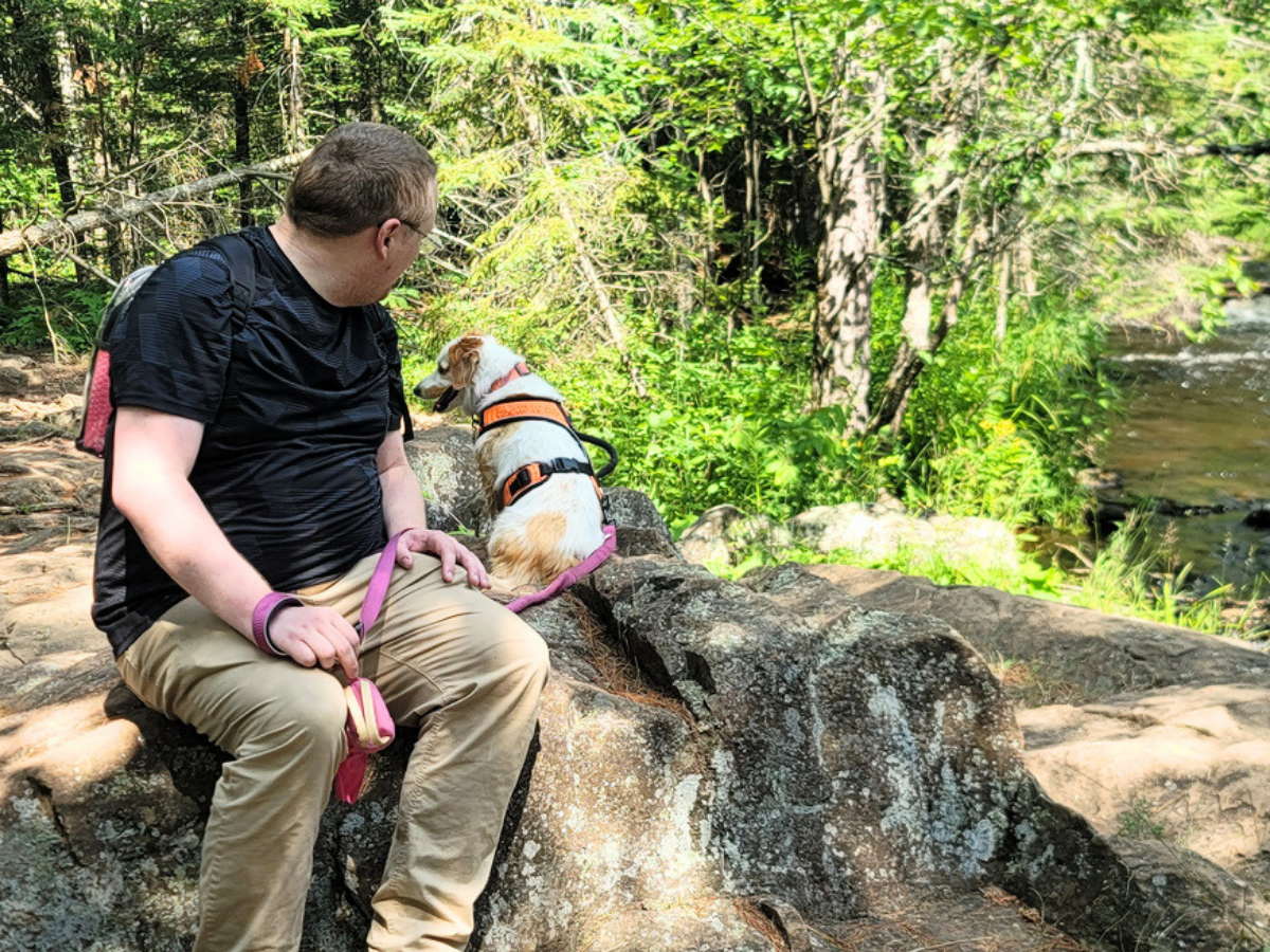 Man in a black shirt and khaki pants sits on a rocky ledge in a sunlit forest, with a small white-and-brown dog wearing an orange harness beside him. A pink leash rests on the rock.