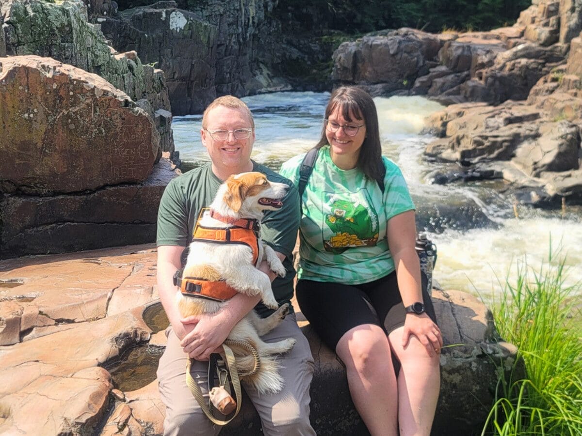 Man holding a small dog in an orange life jacket, sitting on rocks by a rushing river with a smiling woman beside him.