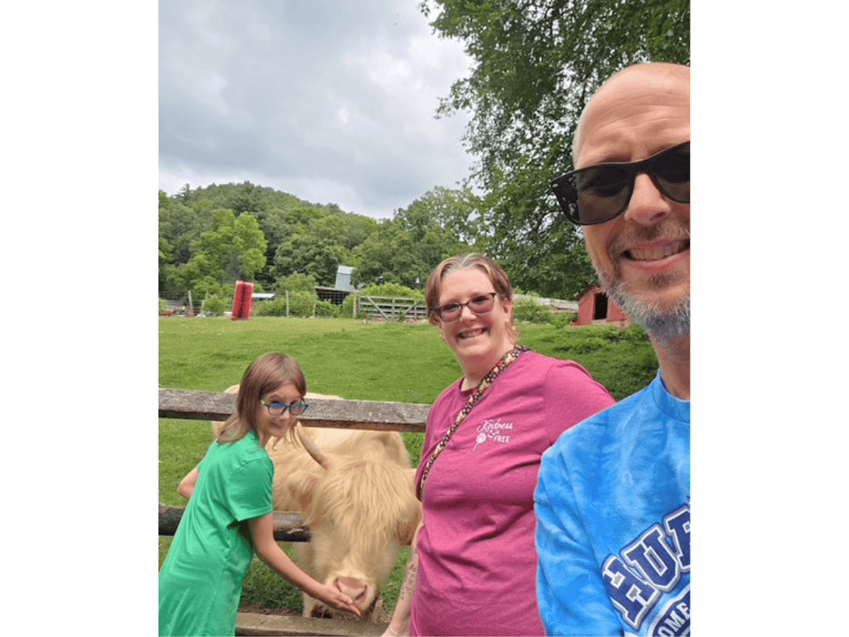 Selfie at a farm with a girl in green, a woman in pink, and a man in sunglasses posing with a cow by a wooden fence.