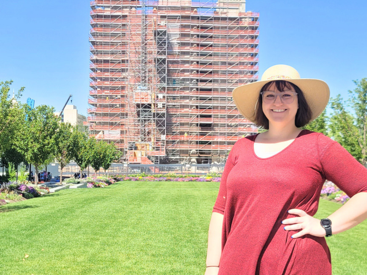 Smiling woman in a red top and wide-brim hat posing on a green lawn with a scaffolding-covered building in the background.