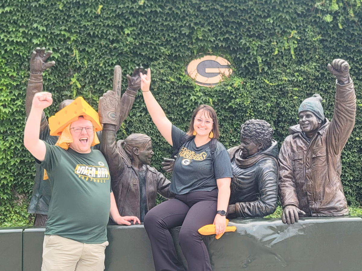 Two Packers fans pose with life-size bronze statues against an ivy wall; man on the left wears a cheesehead and cheers, woman sits on the ledge with a smile.