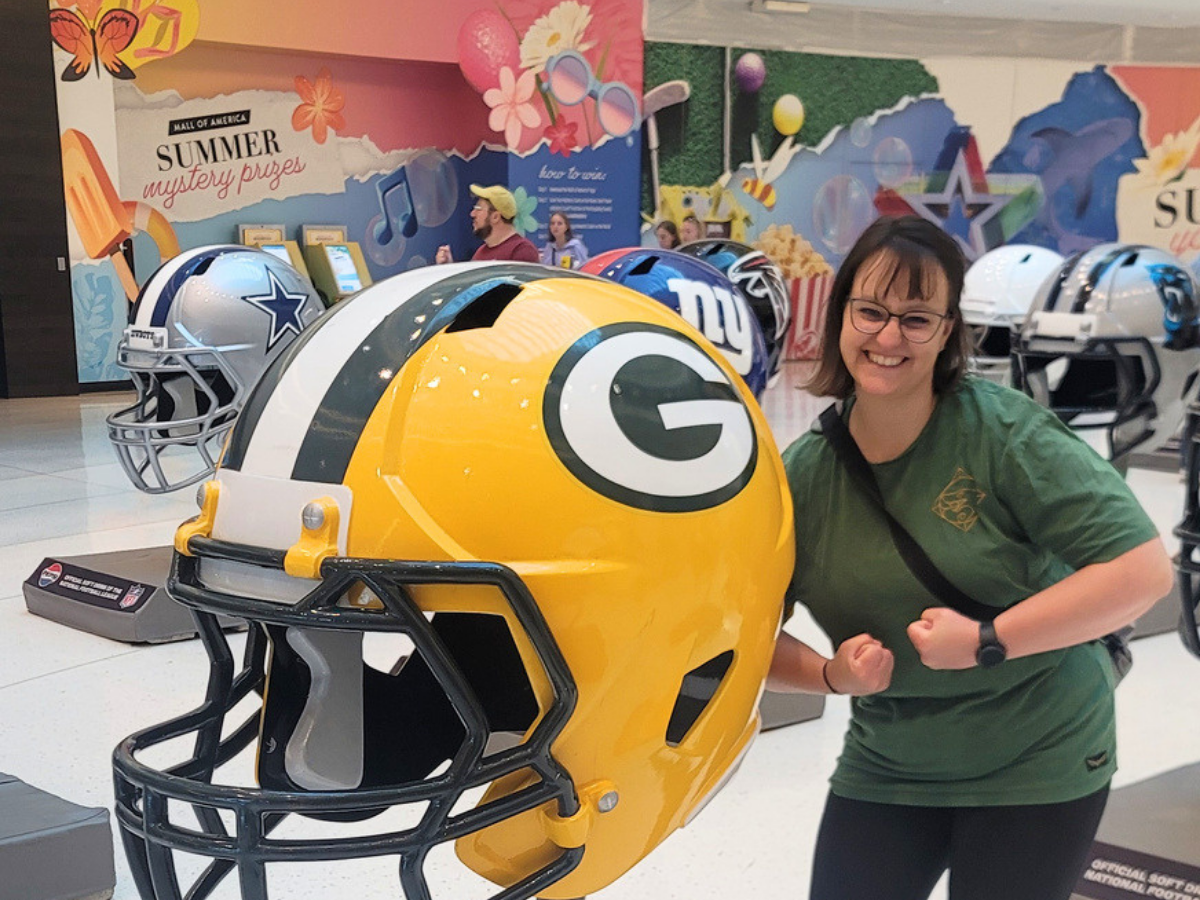 Smiling woman in a green shirt poses with a giant Green Bay Packers football helmet in a bright, colorful display room.