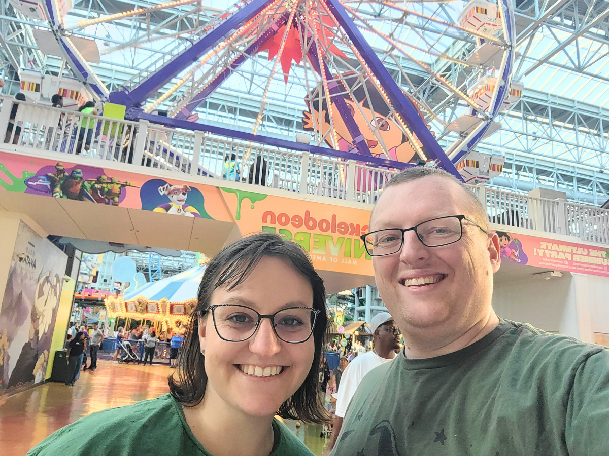 Smiling couple takes a selfie inside a bright indoor amusement park with a large colorful Ferris wheel overhead.