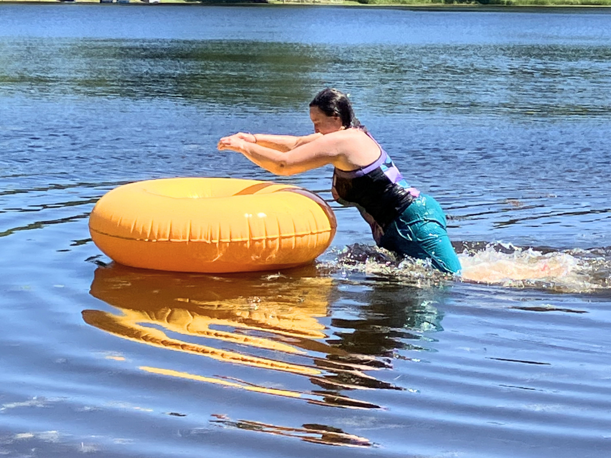 Person wearing a life jacket pushes an orange inflatable inner tube across a calm lake on a sunny day