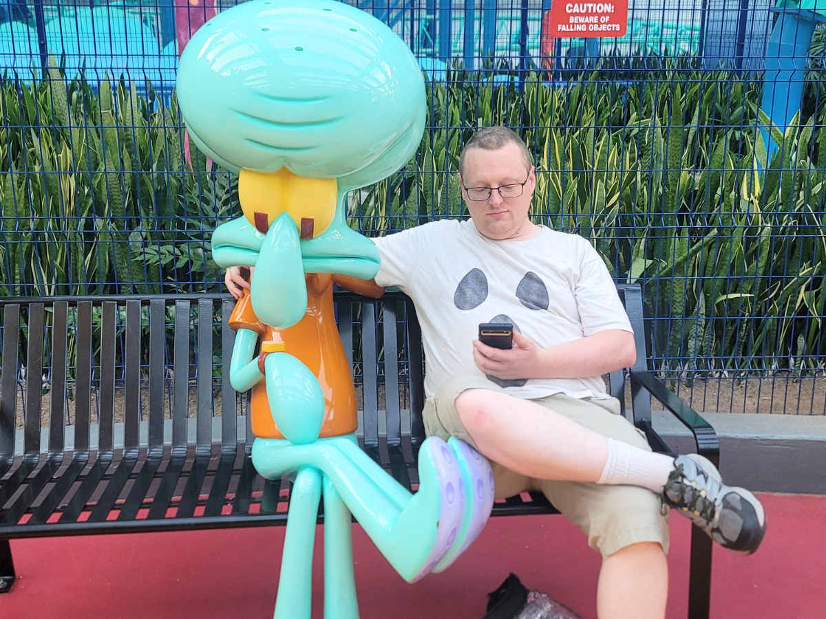 Man sitting on a bench next to a large turquoise Squidward statue, looking at his phone.