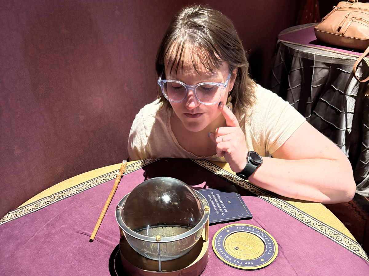 Woman wearing clear-framed glasses leaning over a purple table, gazing into a glass orb on a metal stand as part of a reading setup.