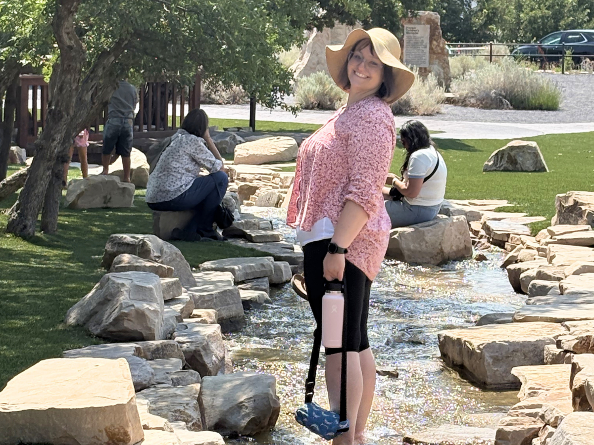 Smiling woman in a wide-brim hat and pink blouse stands in a shallow, rock-lined stream in a park, holding a pink water bottle.