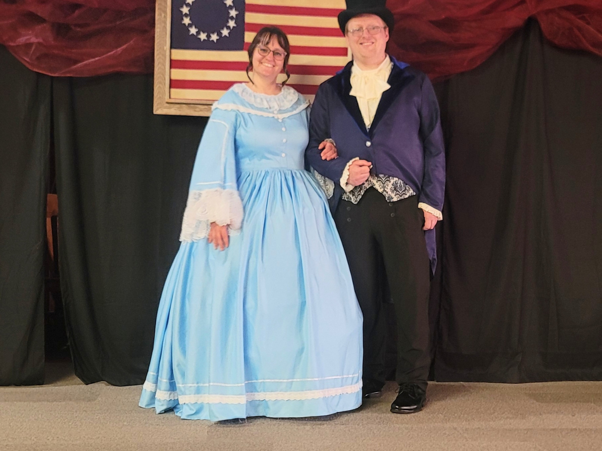 Couple in 19th-century costumes posing on stage: woman in a light blue gown with lace cuffs and the man in a purple tailcoat and top hat, arm in arm in front of a US flag backdrop.