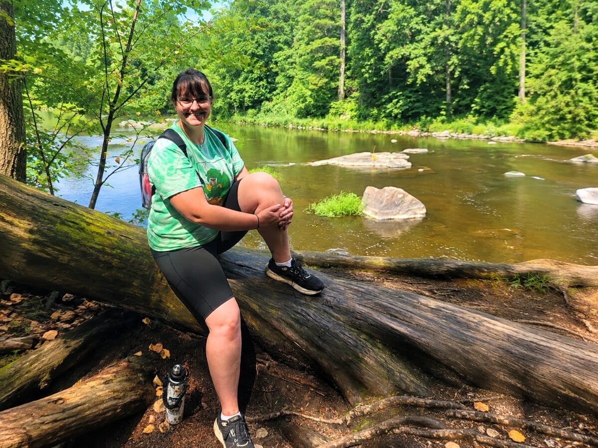 Woman sitting on a fallen log beside a river in a green forest, smiling, wearing a green tie-dye shirt and black shorts with a backpack nearby.