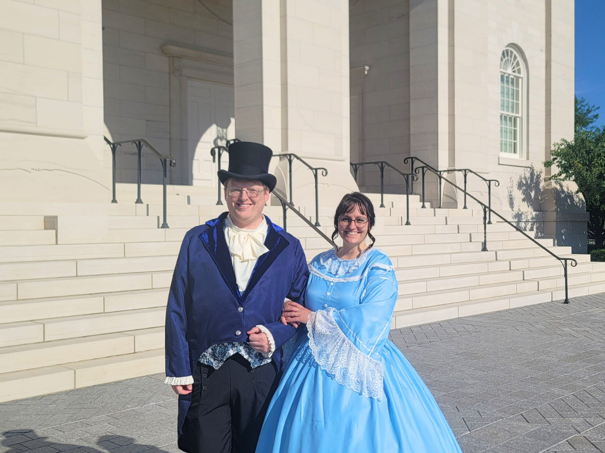 Two people in period costumes pose arm-in-arm on a stone staircase in front of a classical building, smiling at the camera.