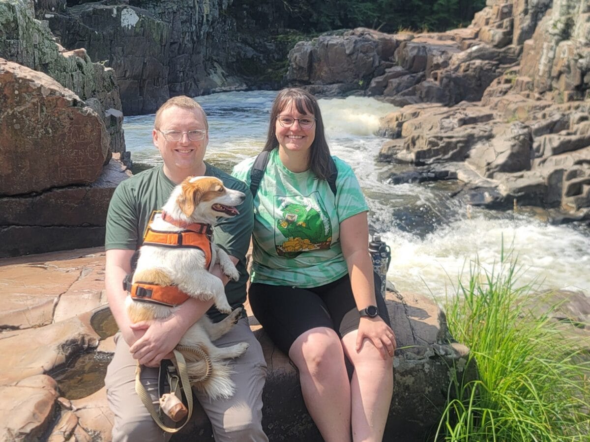 Couple sitting on a rocky riverside with a small dog in an orange life jacket, smiling at the camera near a flowing waterfall.