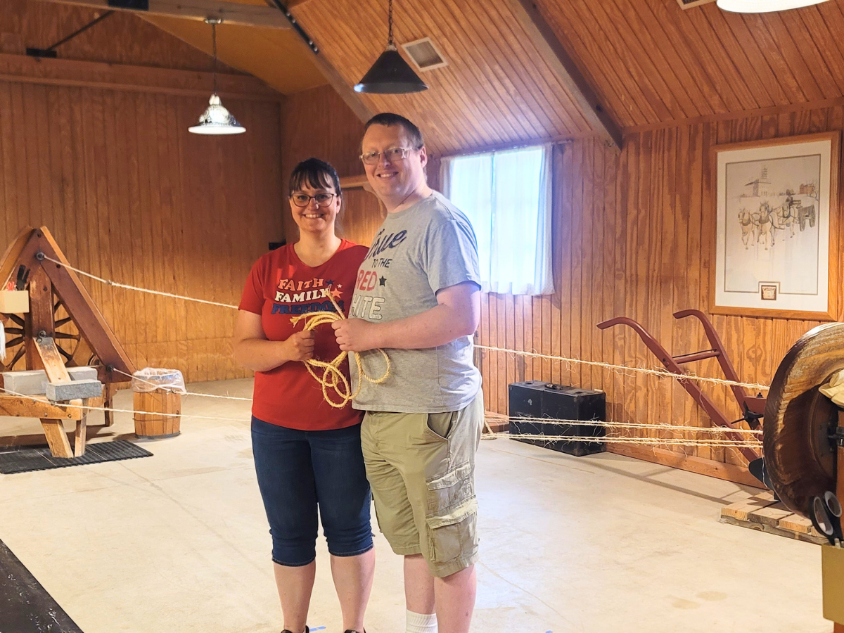 Smiling couple standing in a wooden museum room, holding a rope together in front of vintage equipment.