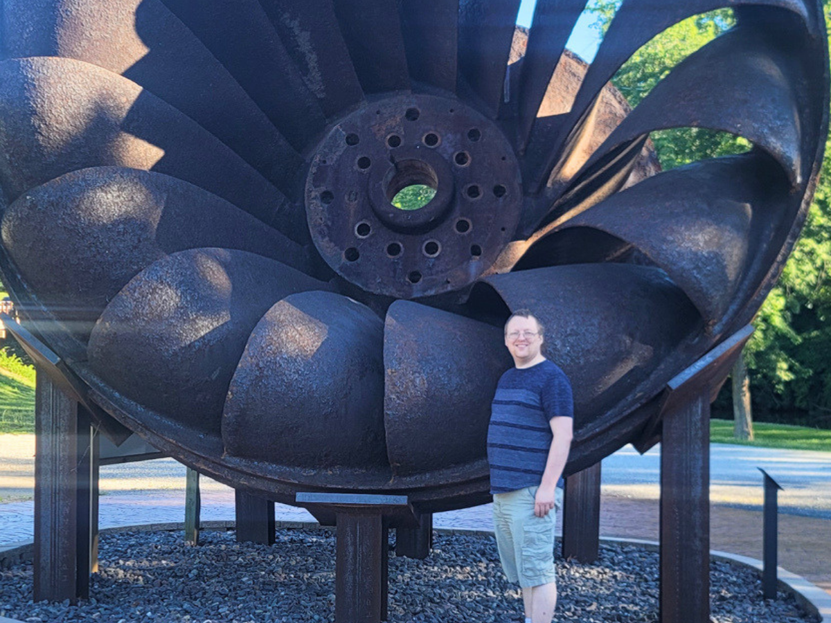 Man standing beside a giant rusted turbine sculpture outdoors in a park, smiling at the camera.