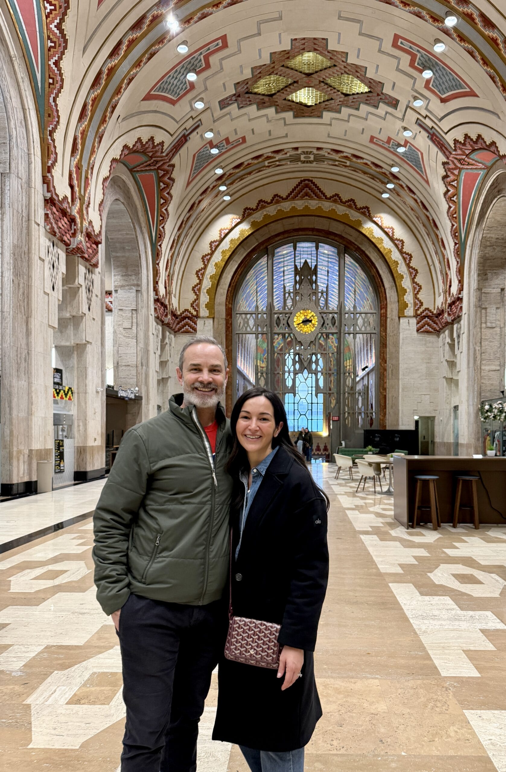Two smiling adults pose in a richly decorated arched hallway with ornate geometric patterns on the ceiling behind them.
