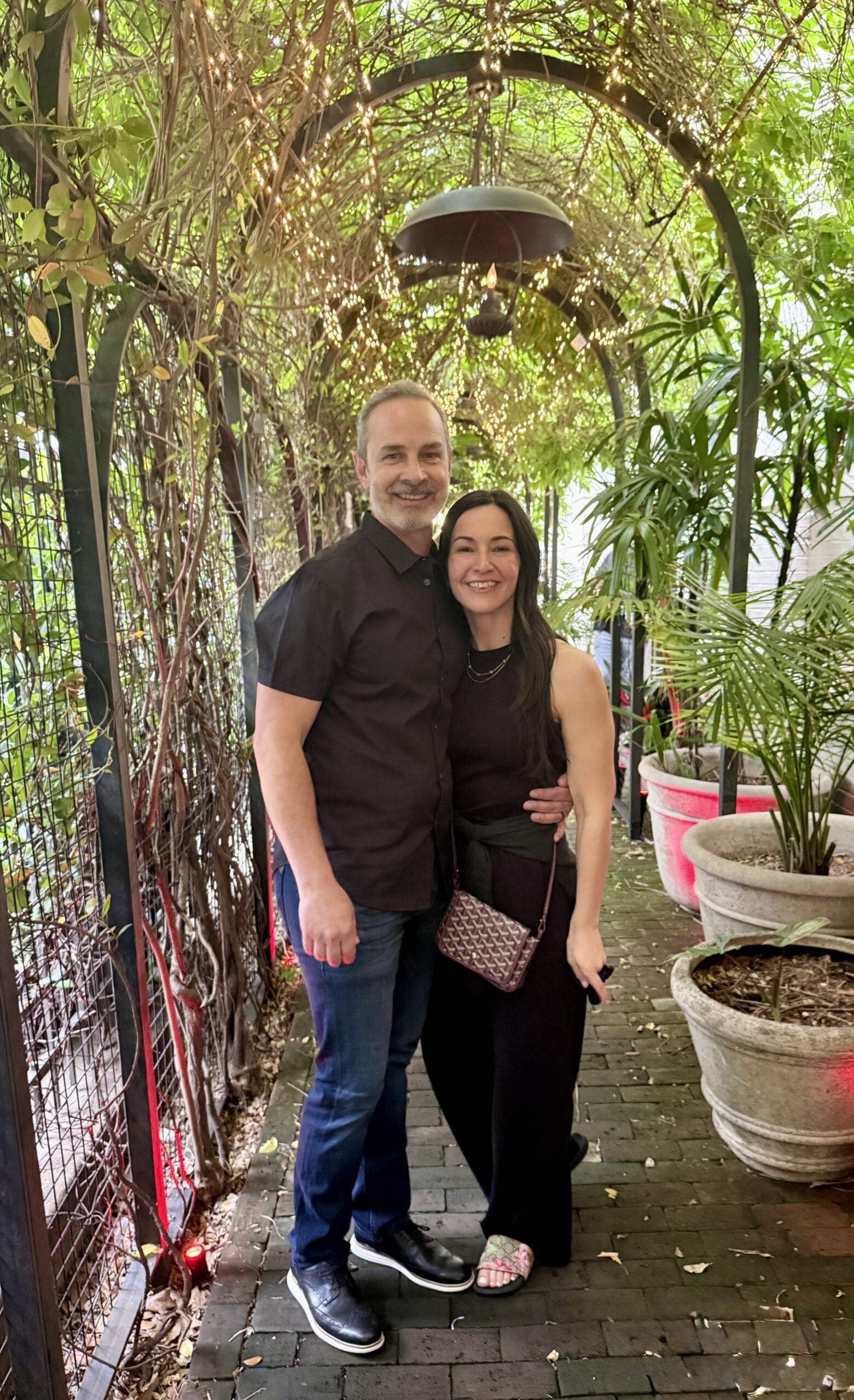 Smiling couple standing together in a vine-covered garden archway with warm string lights overhead.