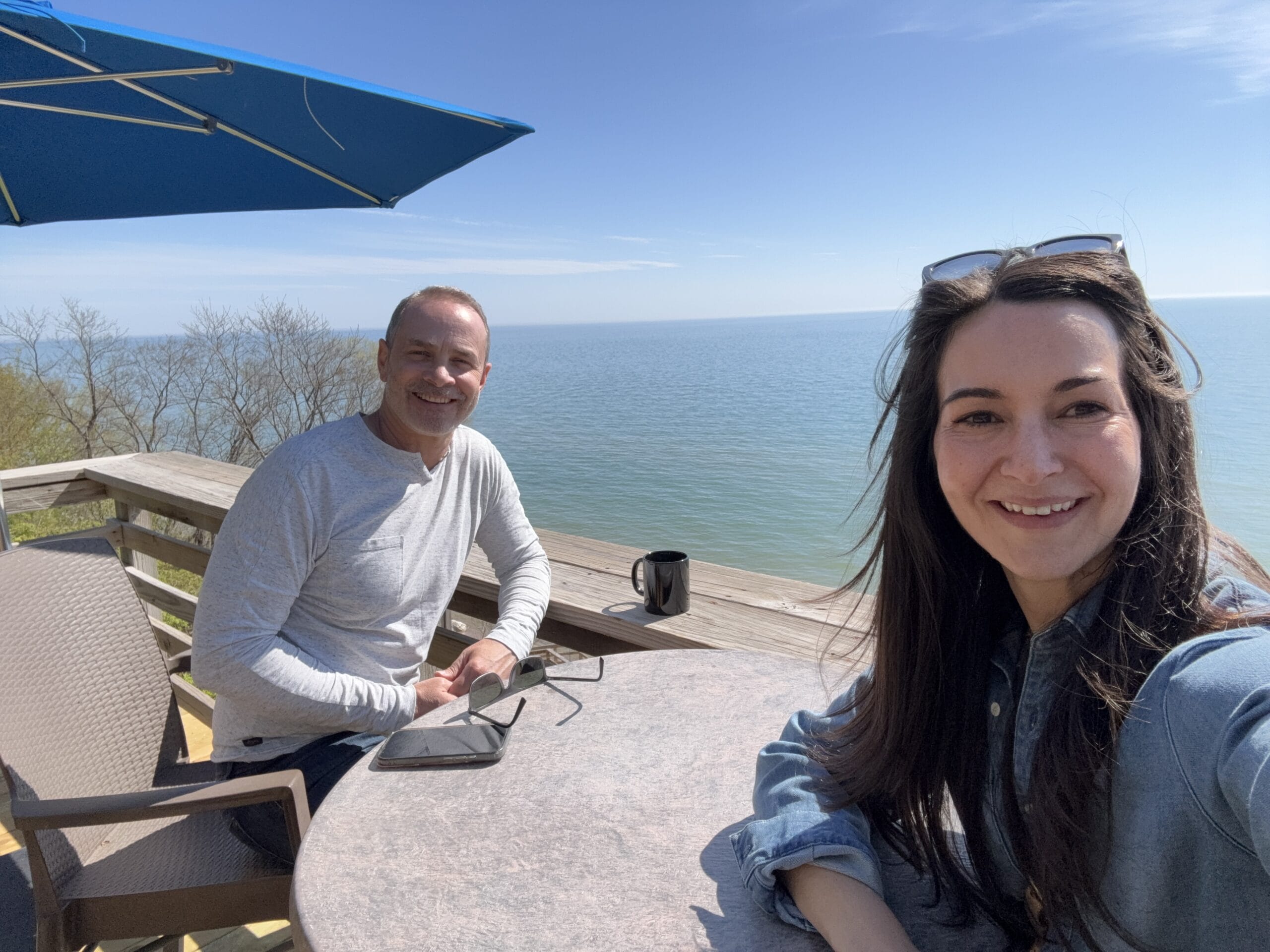 Selfie of a woman in a denim shirt at a seaside outdoor cafe, with a smiling man at the next table and the ocean behind them.