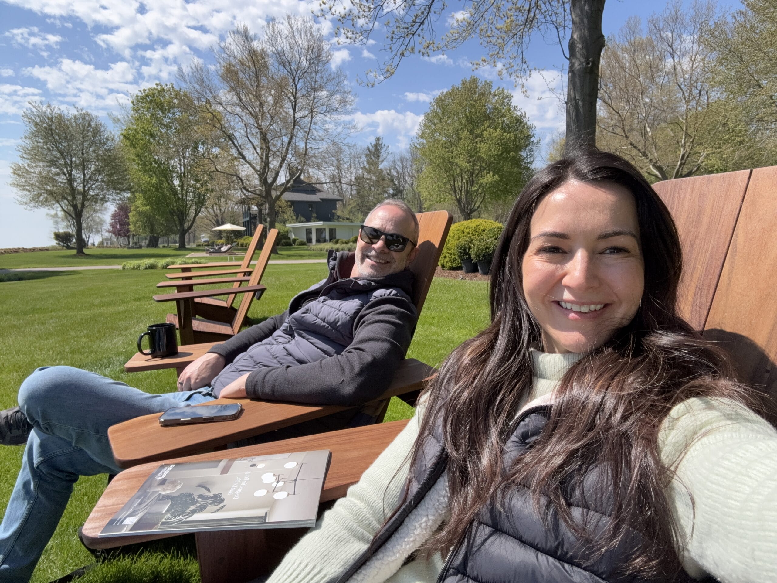 Two people lounging on wooden outdoor chairs in a sunny park; a smiling woman in foreground and a man wearing sunglasses in the background with a magazine on the table.