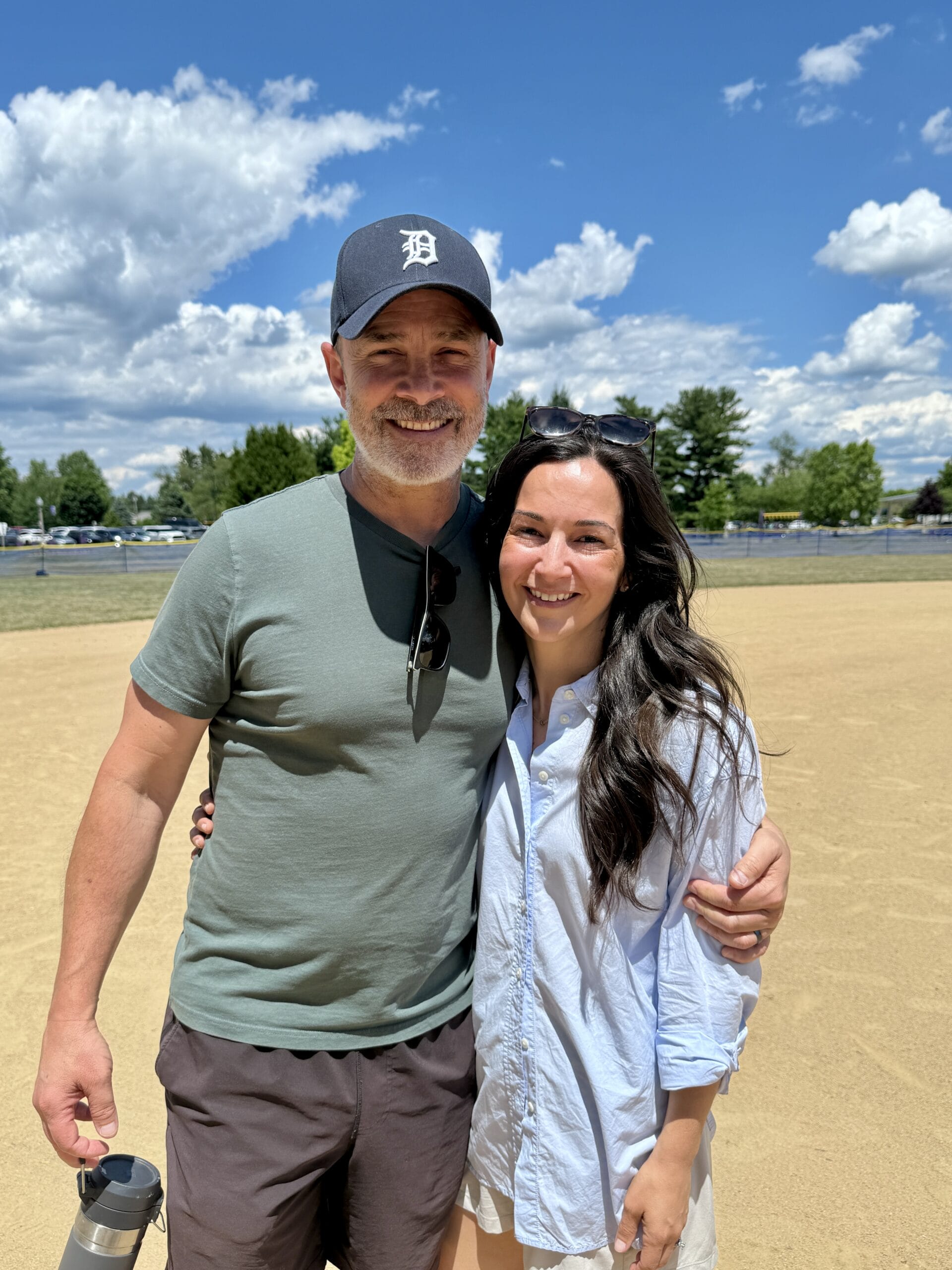 Smiling couple posing outdoors in a sunny park; man in a green t-shirt and a Detroit cap, holding a water bottle, with a woman in a light blue shirt beside him.