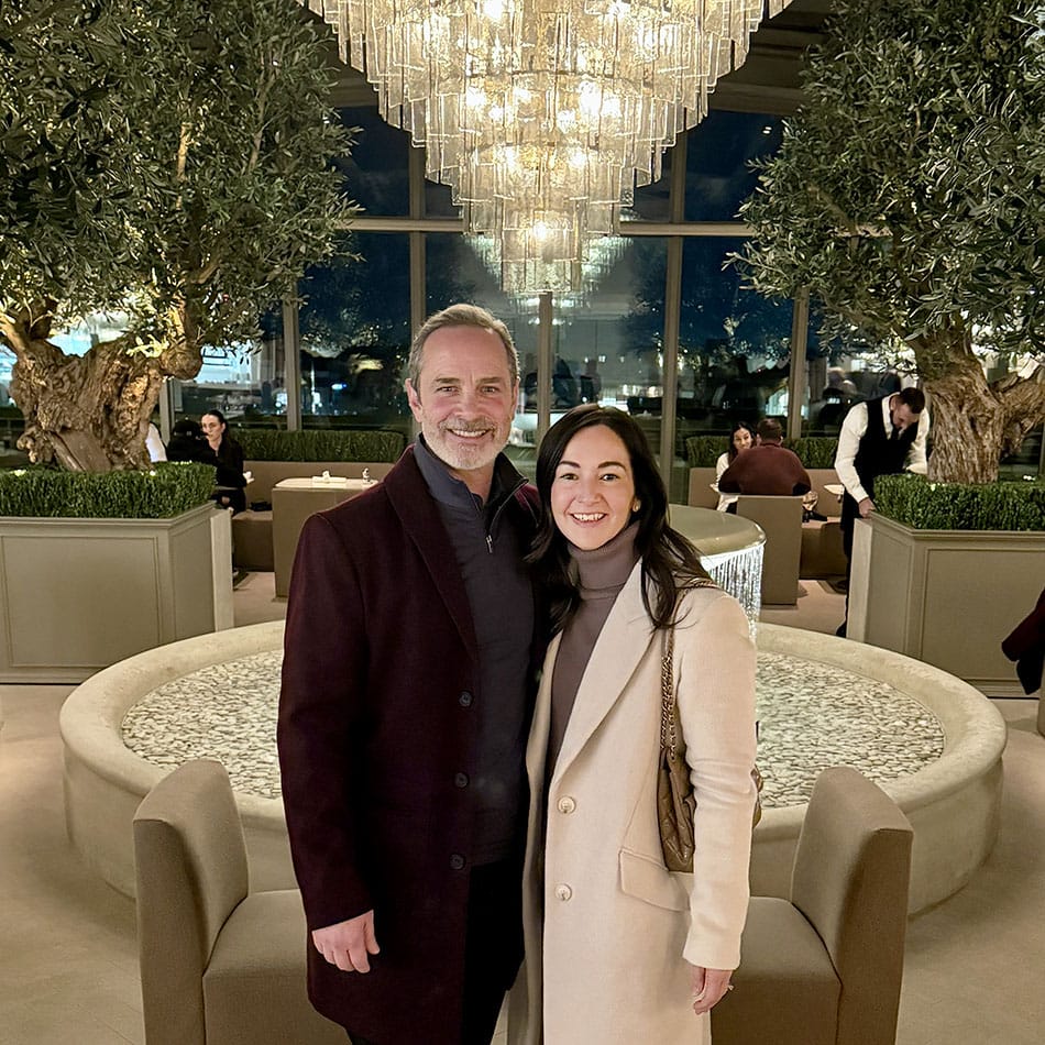 Smiling couple poses together in a luxurious hotel lobby with a large crystal chandelier, circular fountain, and olive trees in planters behind them; man wearing a burgundy coat and the woman in a beige coat.