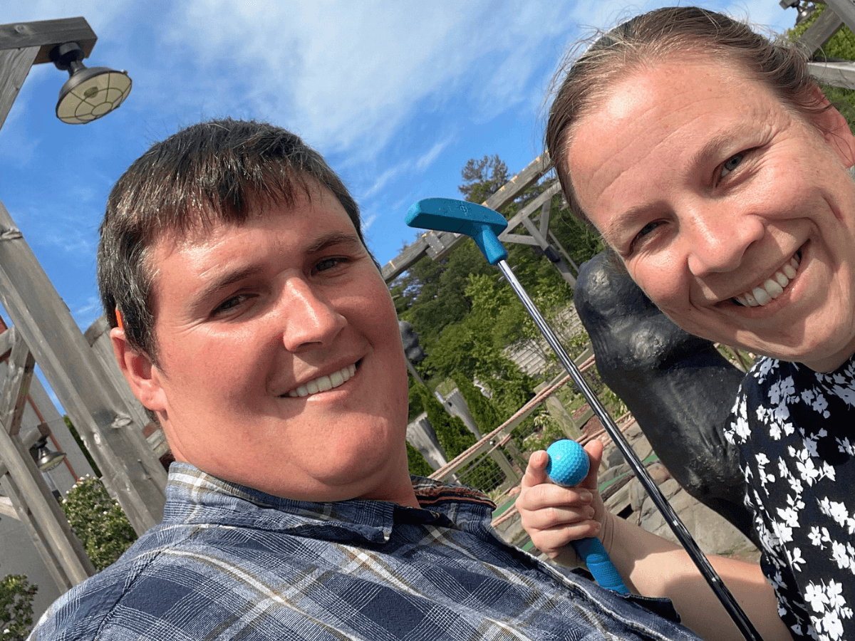 Two friends smile for a sunny outdoor selfie, holding a blue-handled golf mallet and ball near a wooden ride structure.