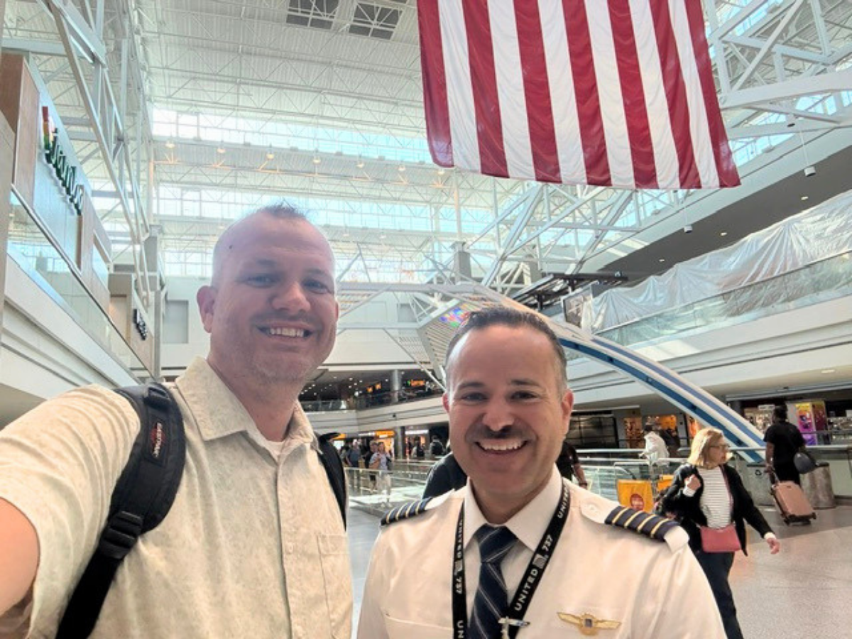 Two men take a selfie in an airport: a casually dressed man with a backpack and a smiling pilot in uniform posing together, with a large American flag overhead.