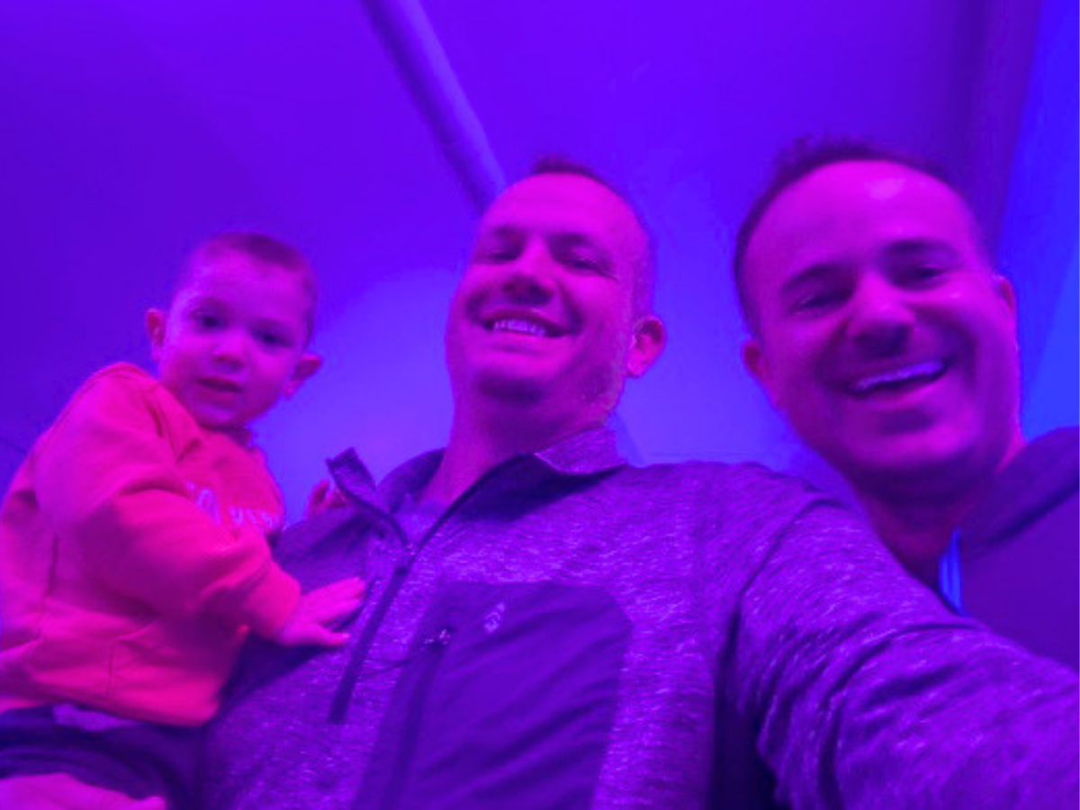 Selfie of two men with a young child, all smiling under blue-purple lighting in a casual setting