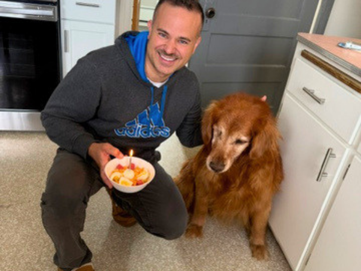 Man kneels in a kitchen holding a bowl with a lit birthday candle while a fluffy brown dog sits beside him on the floor.