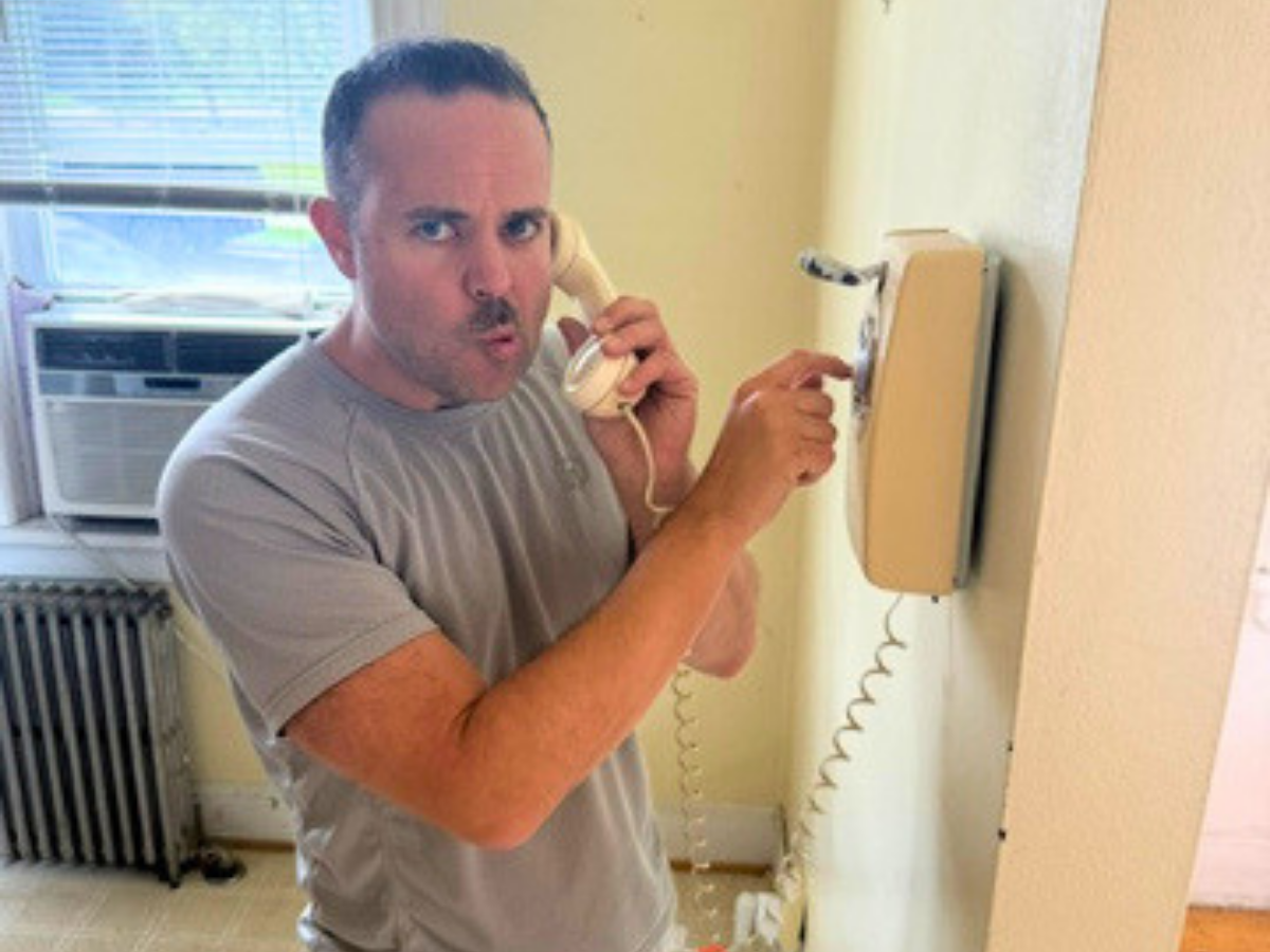 Man in a gray t-shirt talks into a wall-mounted beige landline, making a surprised pursed-mouth expression in a sunlit room with an air conditioner behind him.