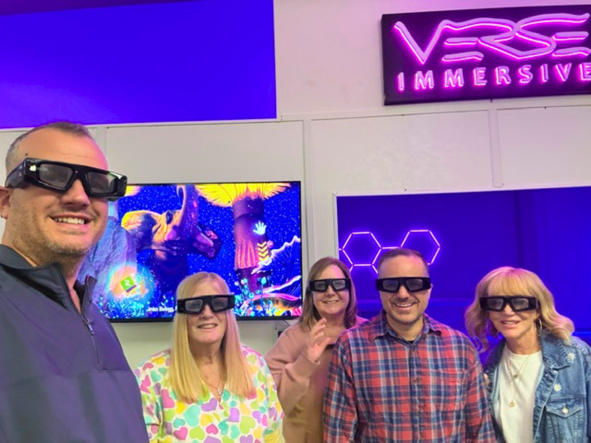 Five adults wearing virtual reality headsets pose for a photo in a neon-lit VR showroom with a Verse Immersive sign in the background, smiling at the camera, colorful screens behind them.