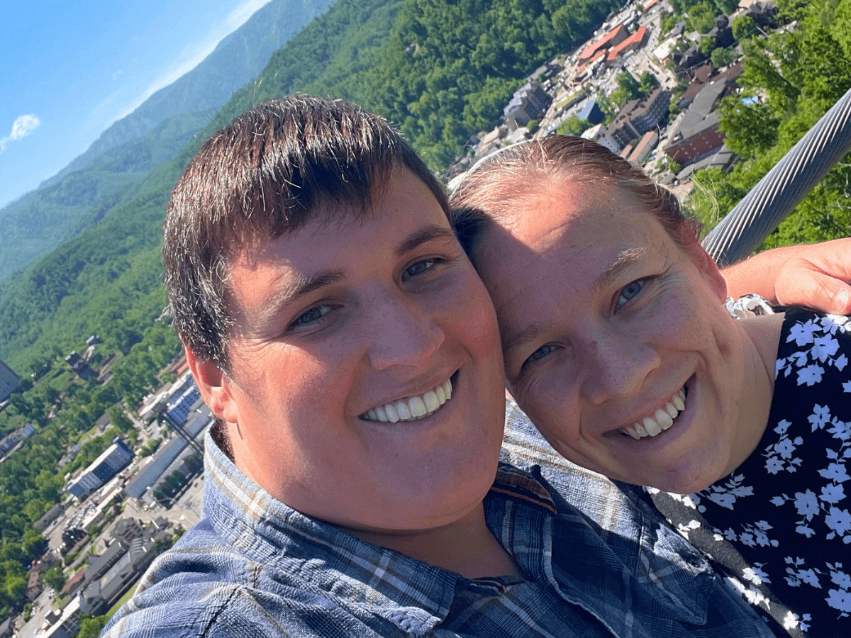 Two friends take a sunny selfie with a green, mountainous town in the background, smiling at the camera.