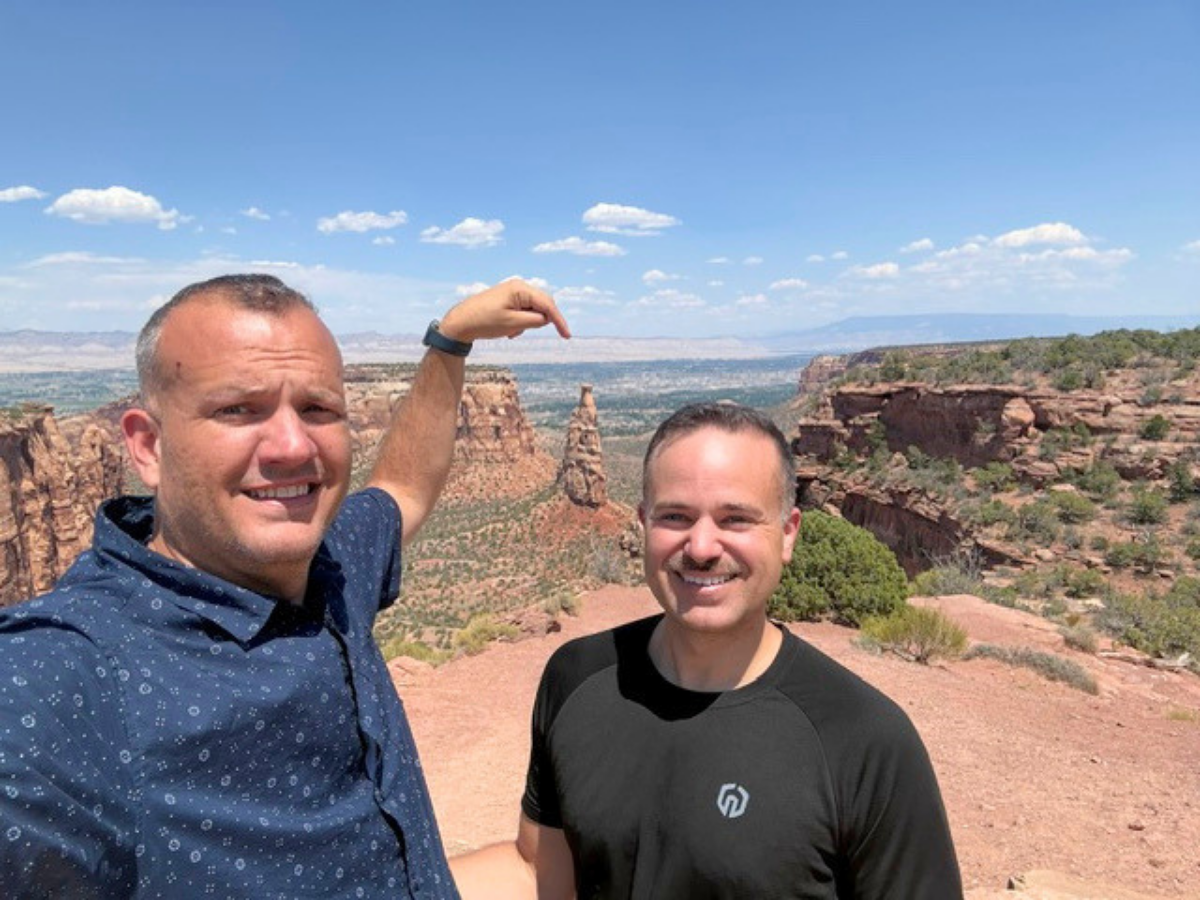 Two men posing on a red rock overlook in a desert canyon beneath a blue sky, one pointing toward distant rock formations, smiling.