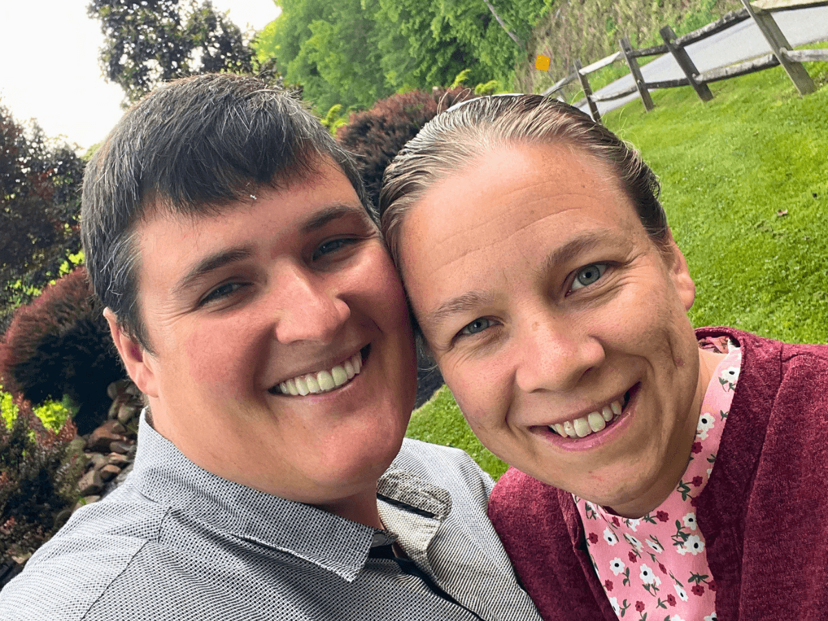 Two people smiling at the camera, leaning heads together in a sunny outdoor garden?