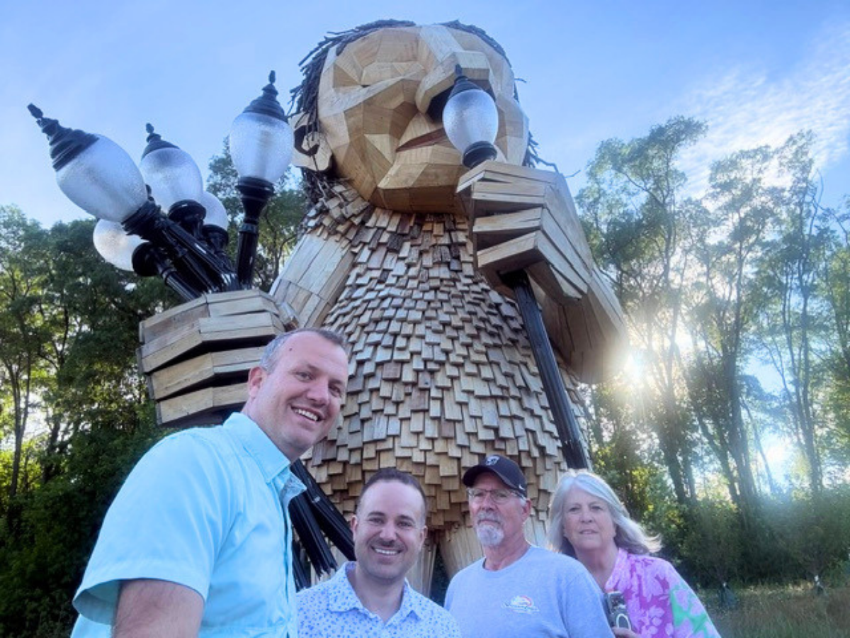 Four people posing in front of a huge wooden humanoid sculpture with lamp posts in a park behind them, smiling for the camera.