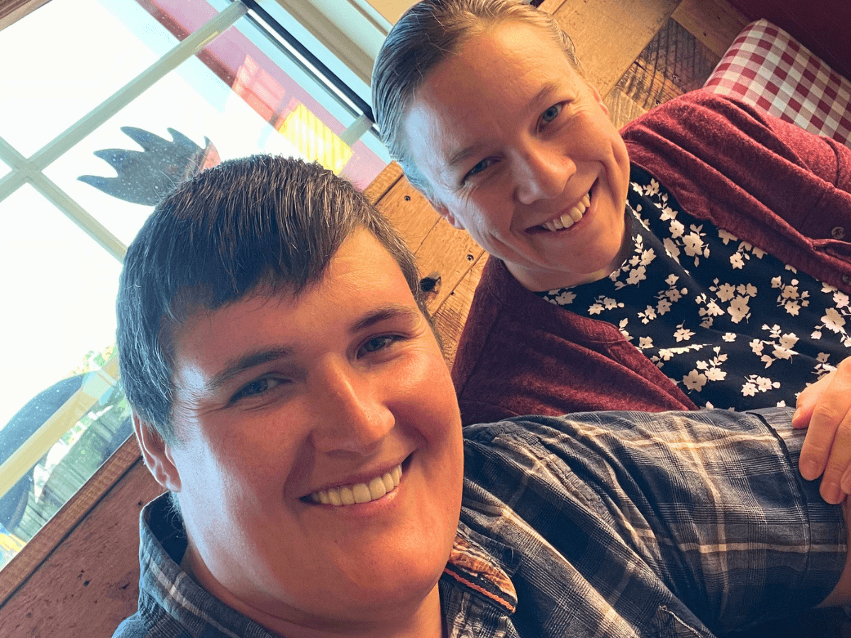 Two friends smile for a selfie indoors, with a wooden wall and window behind them.