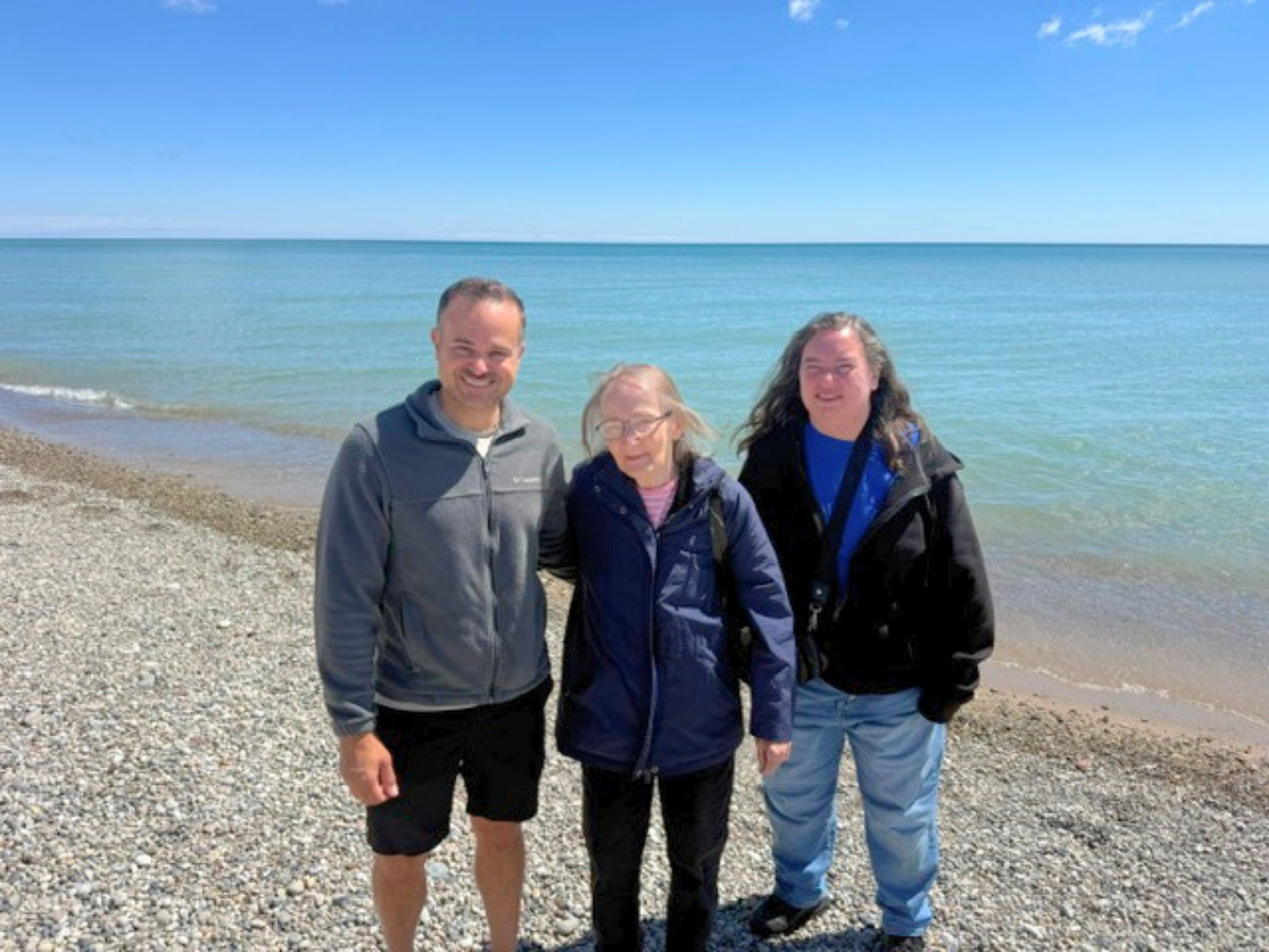 Three people stand together on a rocky beach in jackets, with a calm blue sea and clear sky behind them.