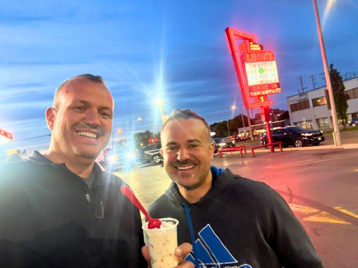 Two men smiling in a parking lot at dusk, one holding a vanilla ice cream sundae with a cherry on top behind them.