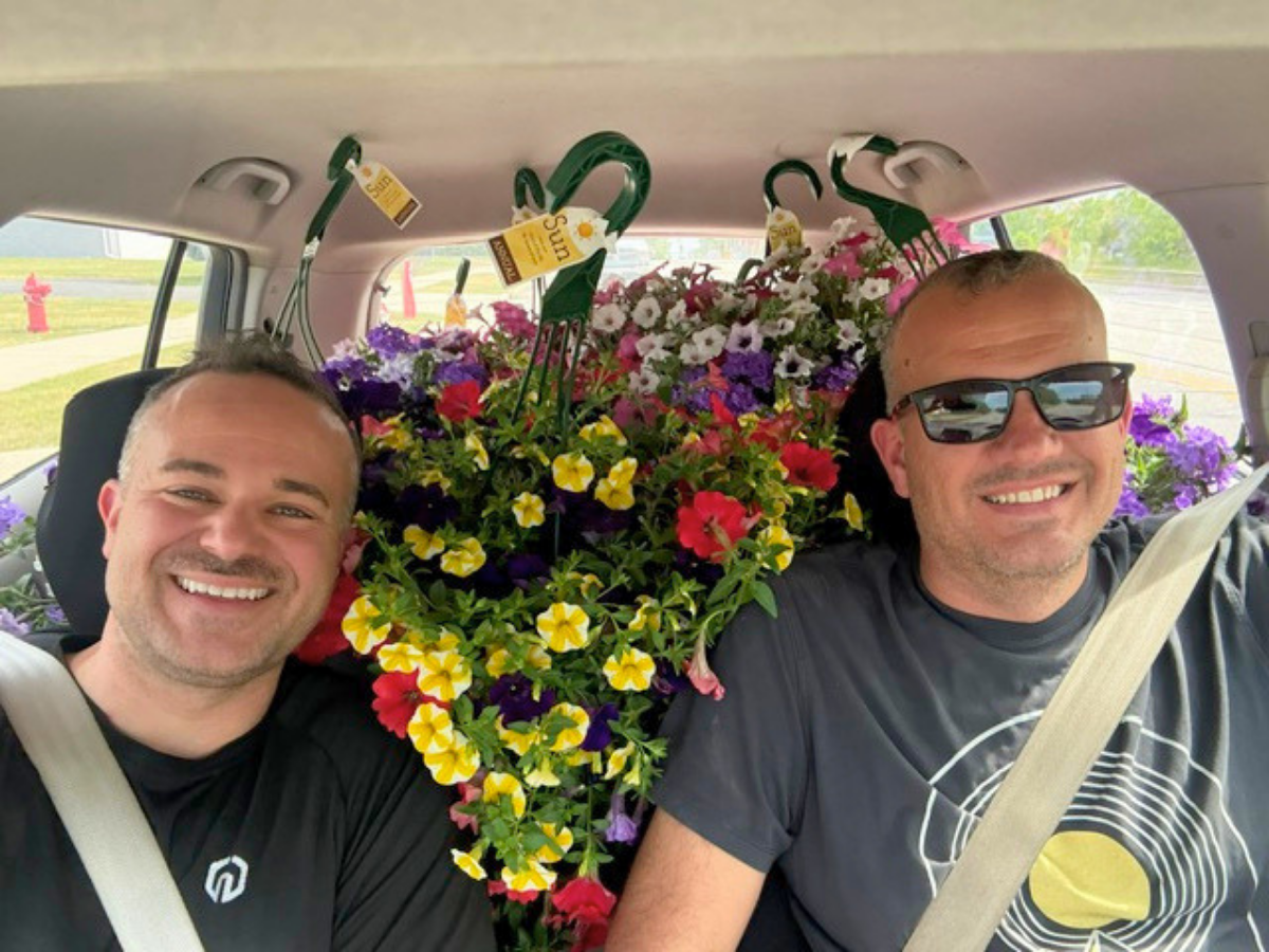 Two smiling men in a car wearing seatbelts with a large colorful flower arrangement between them on the back seat area.