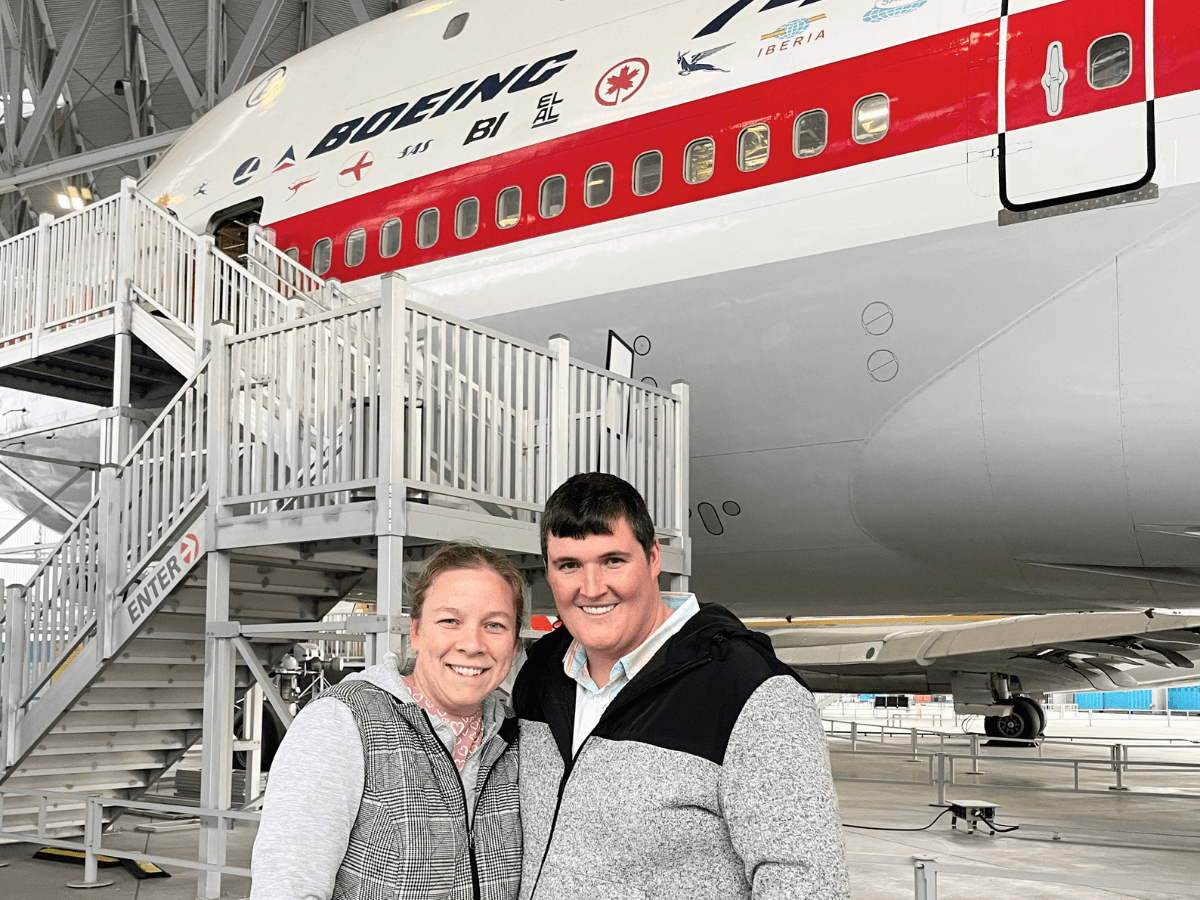 Two smiling people pose in front of a large Boeing airplane inside a maintenance hangar with a mobile staircase nearby.