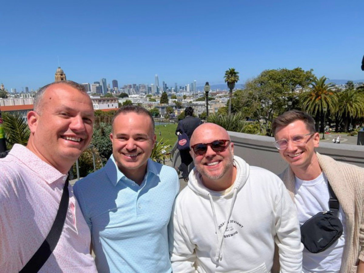 Four men smiling for a selfie on a sunny terrace with a city skyline in the background.