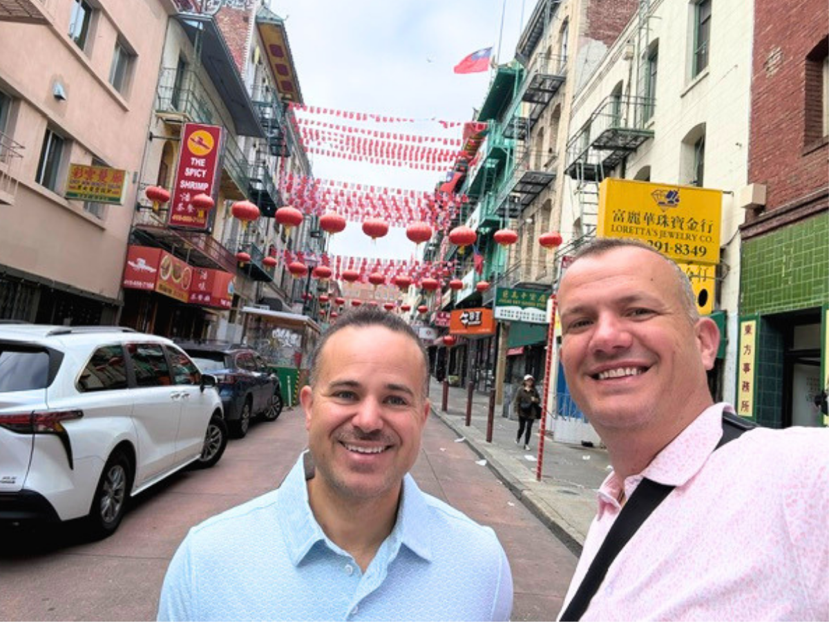 Two men smiling as they take a selfie on a Chinatown street lined with red lanterns and colorful storefronts.