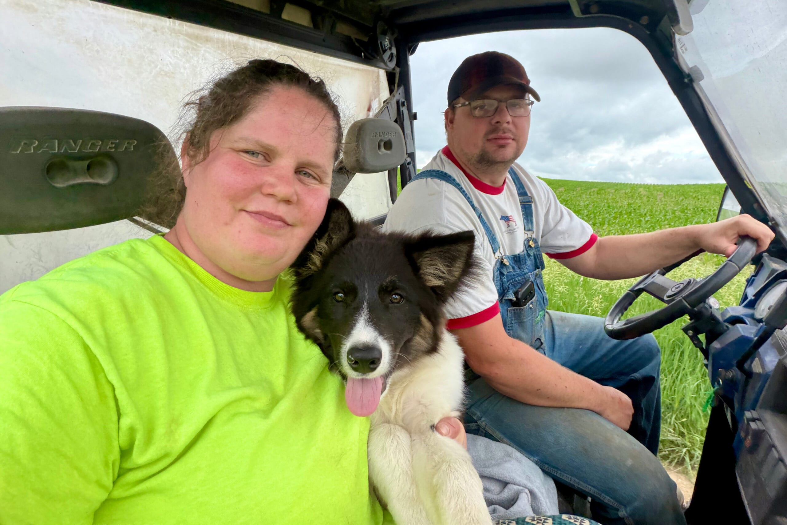 A black and white puppy sits on a person’s lap in a lime green shirt as two men ride in a truck across a green field with a cloudy sky.