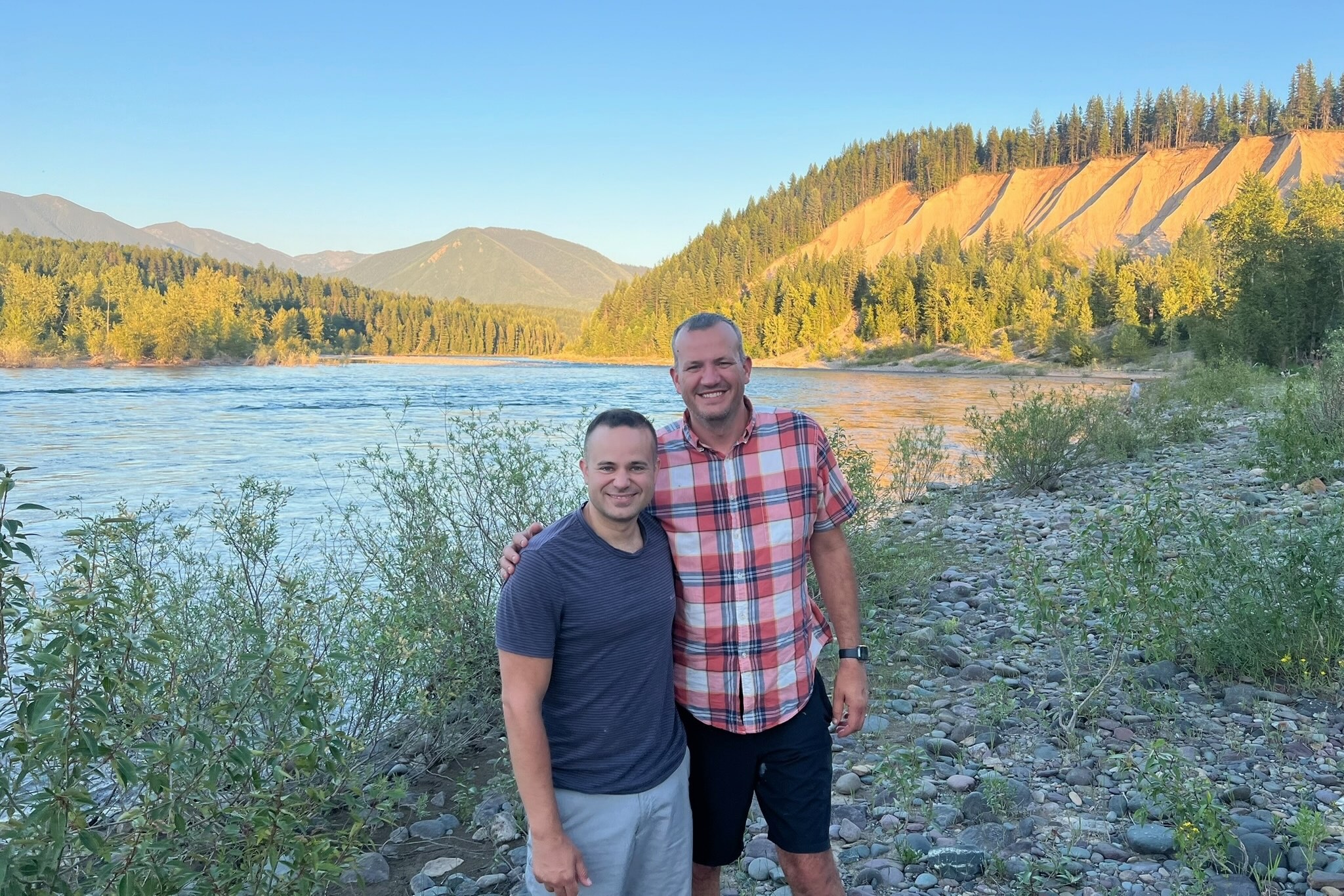 Two men stand together on a rocky riverbank, smiling, with a forested hillside and blue sky in the background.