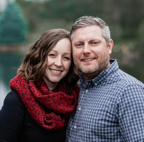 Smiling couple posing closely outdoors by a lake; woman wears a red scarf, man in a blue plaid shirt.