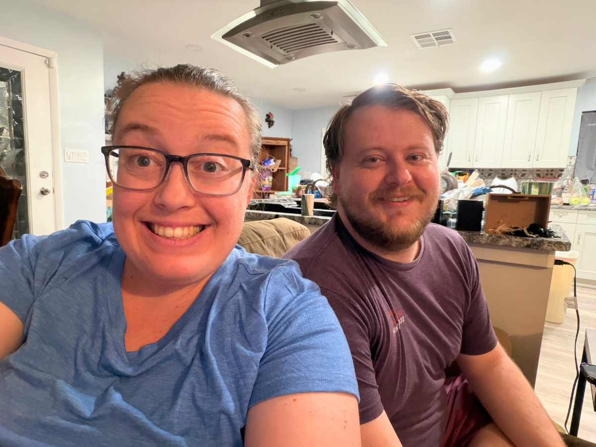 Two adults sit on a couch in a bright kitchen, smiling at the camera; cluttered countertops and cabinets behind them.