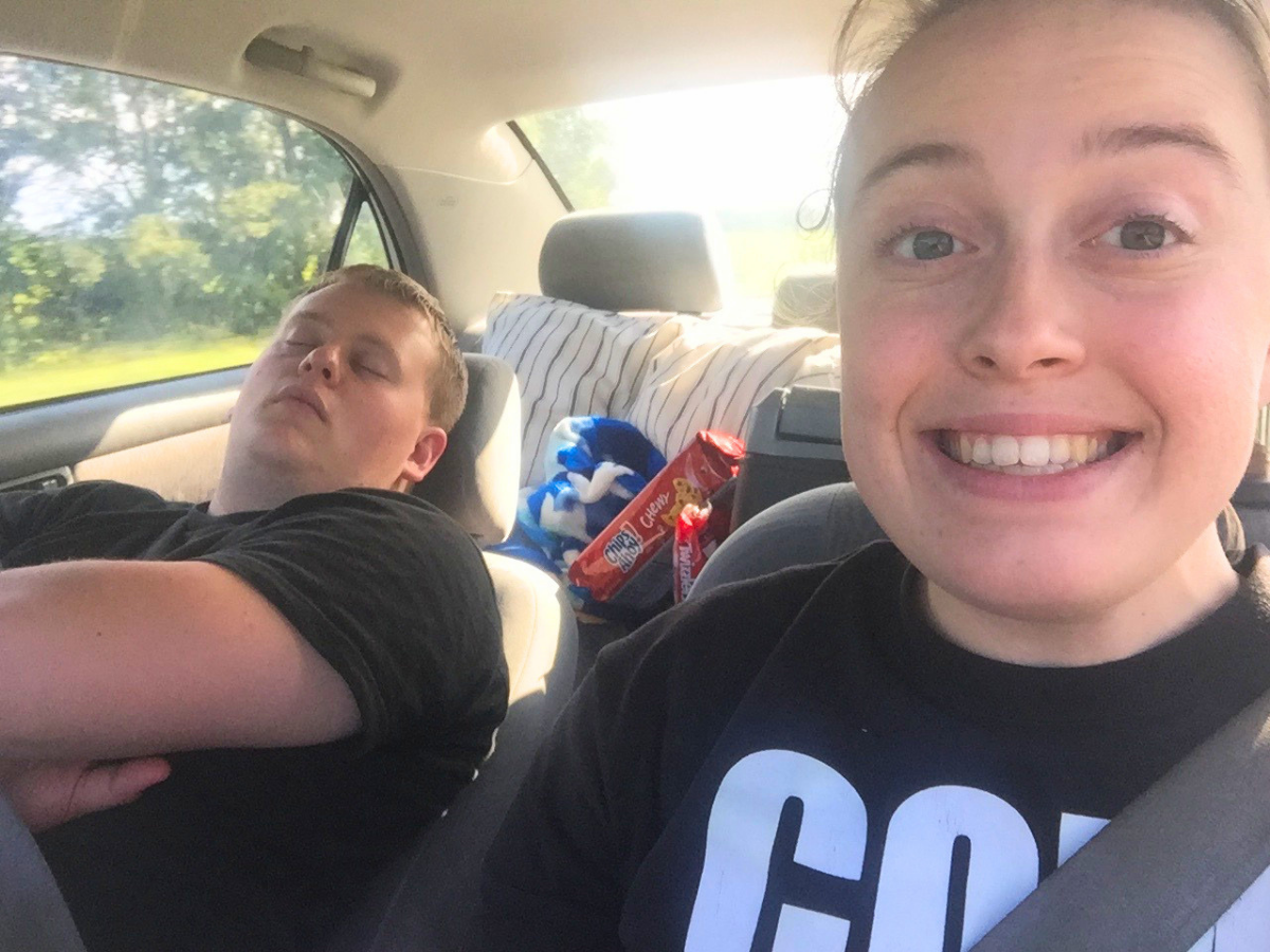 A woman in the front passenger seat smiles at the camera while a man in the back sleeps, with snack bags visible behind them inside the car.