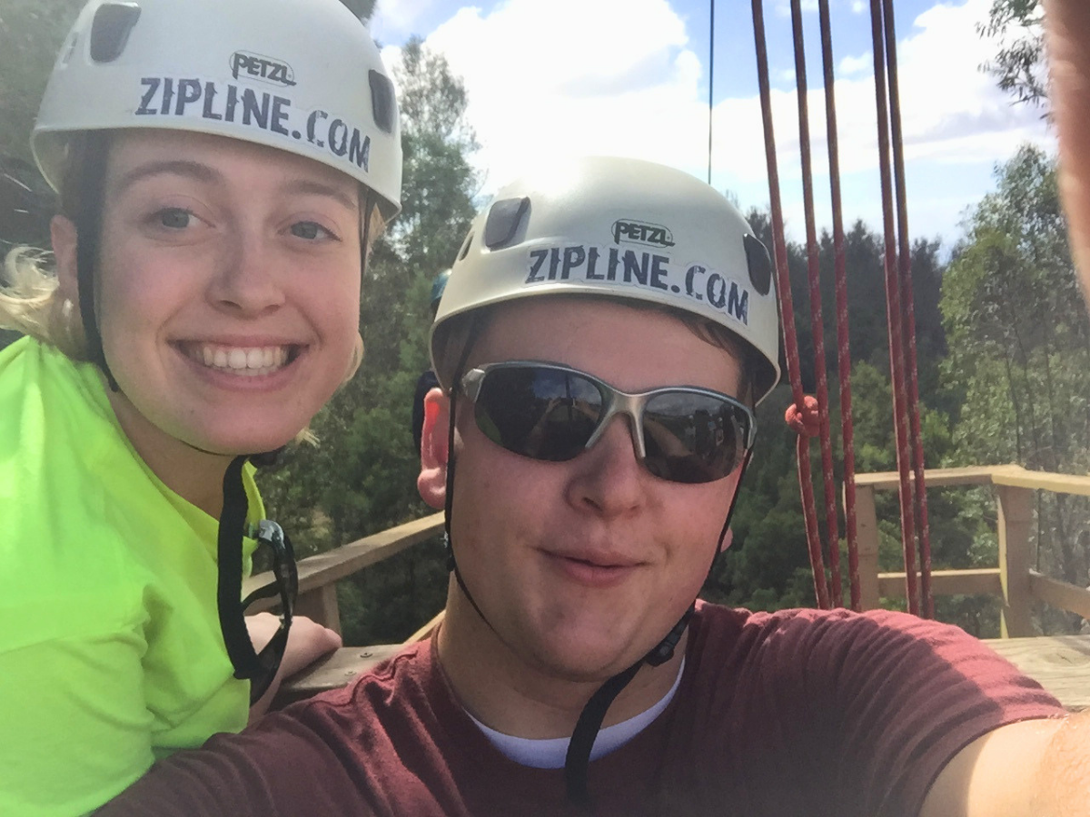 Two friends wearing helmets labeled ZIPLINE.COM pose for a selfie on a wooden zipline platform outdoors, smiling at the camera.