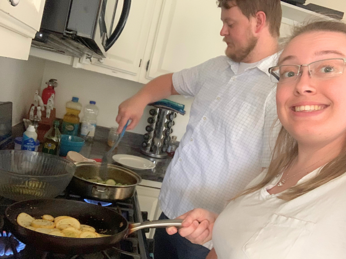 Smiling woman taking a selfie in a kitchen as a man stirs food in a pan on the stove.