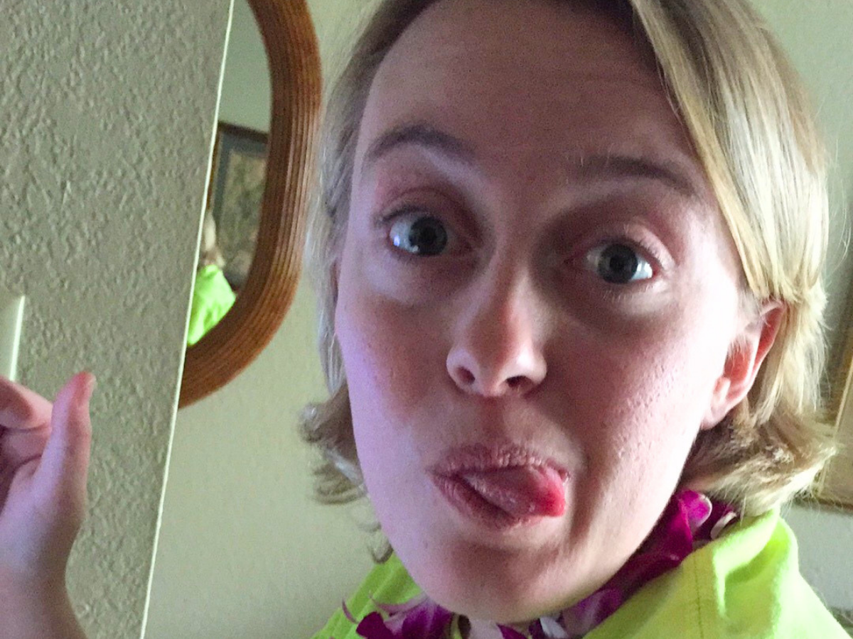Person with blonde hair making a goofy face, eyes wide and tongue sticking out, wearing a neon scarf indoors near a mirror.