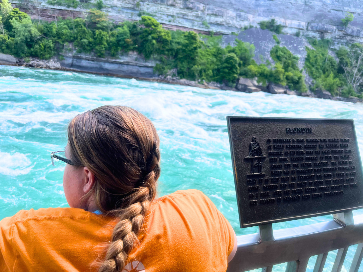 Person with a braided ponytail in an orange shirt leans on a railing, watching turquoise river rapids; a metal plaque labeled Blondin is to the right.