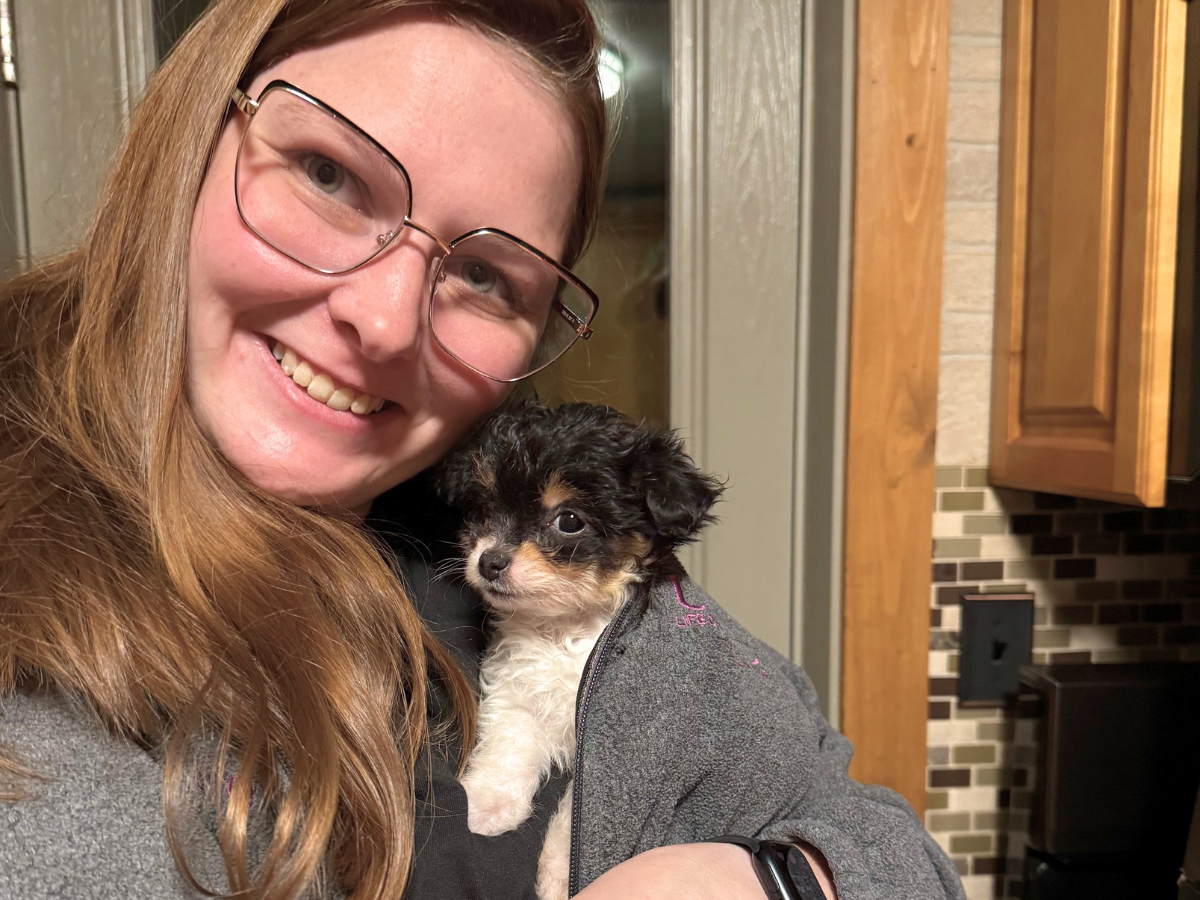 Smiling woman with glasses holding a small black-and-white puppy indoors in a kitchen setting.